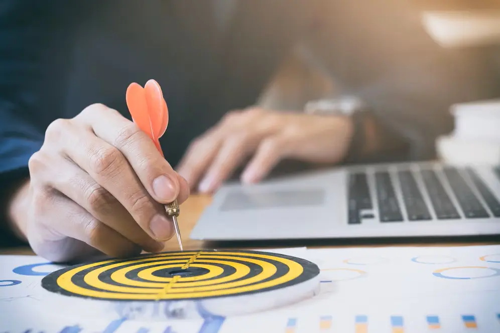 A person places a red dart into the center of a yellow and black target on a desk with charts, near an open laptop used for blogging on WordPress.