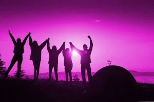 Silhouetted group of five people holding hands and raising them in the air at sunset with a tent and mountain landscape in the background, set against a purple-pink hue. The scene evokes a digital marketing campaign aimed at capturing unity and adventure.