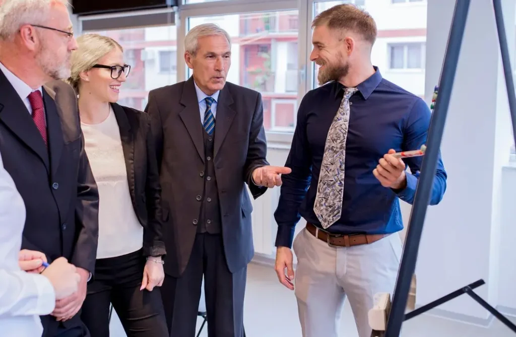 A group of four colleagues in business attire stands near a flip chart, engaged in a discussion about effective marketing strategies in a modern office setting. The conversation touches on opportunities presented by the new Green Freeport initiative.