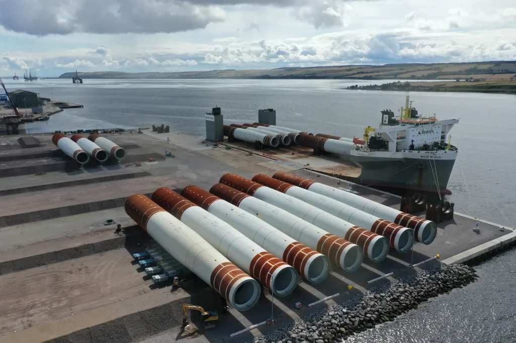 A docked cargo ship is loaded with large wind turbine components, embodying the spirit of a Green Freeport, while additional components are lined up on the pier next to it on a cloudy day.
