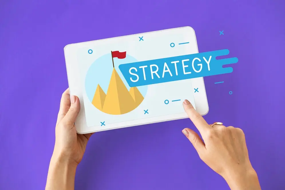 Hands holding a tablet displaying a mountain with a flag, surrounded by the word "Strategy," set against a purple background. Perfect for a marketing agency in Inverness dedicated to reaching new heights.
