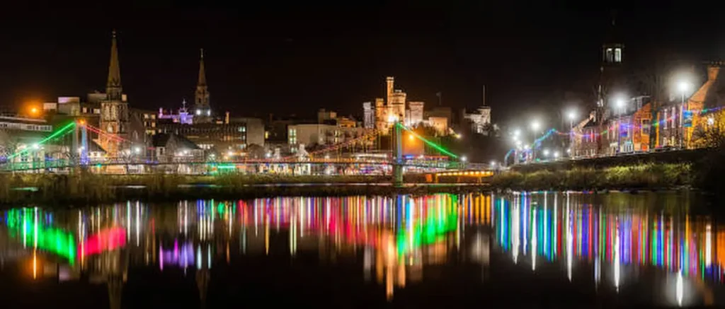 A cityscape at night showcases Inverness with buildings and churches lit up, their colorful reflections shimmering on a river, much like a marketing agency illuminating fresh ideas onto the canvas of commerce.