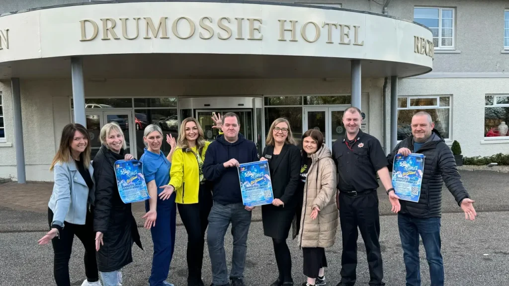 A group of people from a local marketing agency stand smiling in front of the Drumossie Hotel entrance in Inverness, holding event posters.